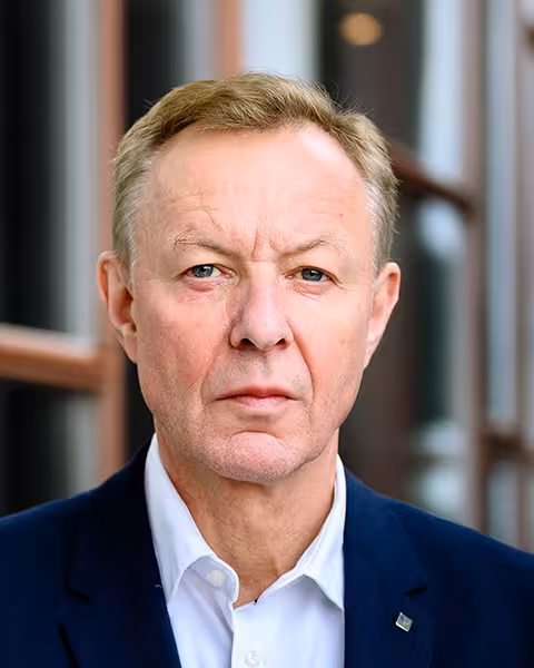 Mann i blå dress med hvit skjorte står utenfor glassvegg og ser i kamera med hendene foldet foran seg.
