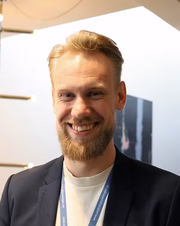 Blond mann med skjegg ser inn i kamera og smiler, mens han står foran store moderne lysekroner.