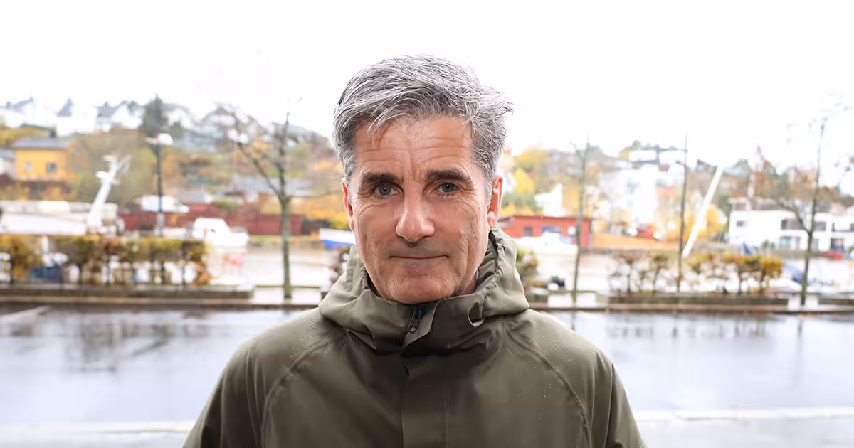 Mann i grønn regnkåpe står på andre siden av veien fra elv og ser inn i kamera.