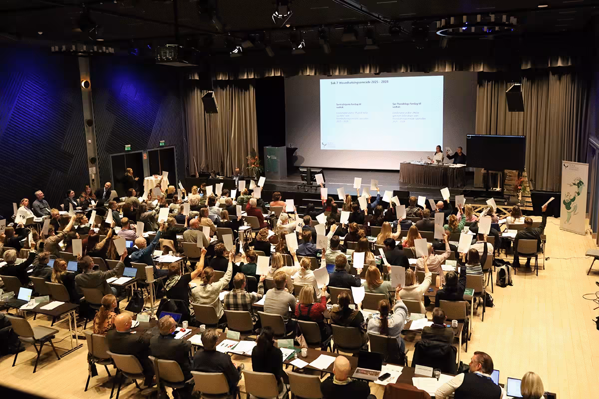 Mennesker som sitter på rader i en sal. De fleste holde oppe hvert sitt hvite ark.