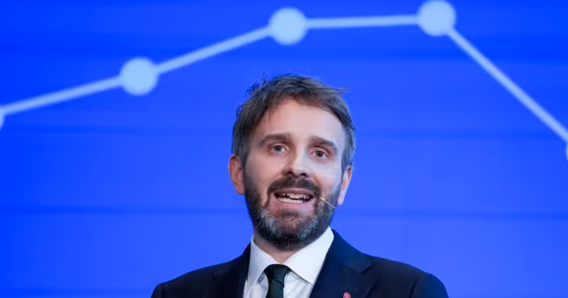 Talende mann i dress med mikrofon, som står foran skjerm og presenterer.