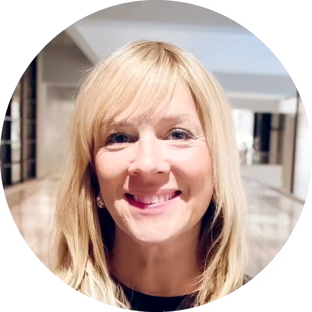 Portrettbilde av blond kvinne i sort genser som står i en passasje, smiler og ser inn i kameraet