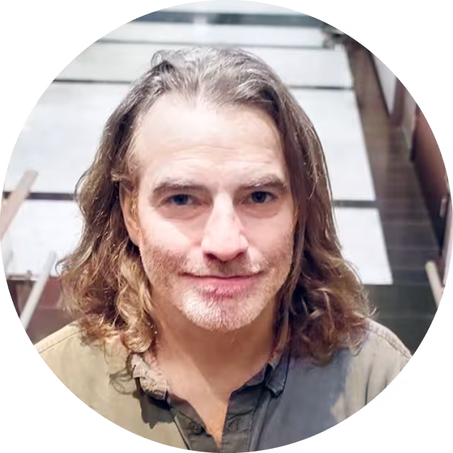 Portrettbilde av mann i skjorte, med langt hår, som står i en trapp, smiler og ser inn i kameraet