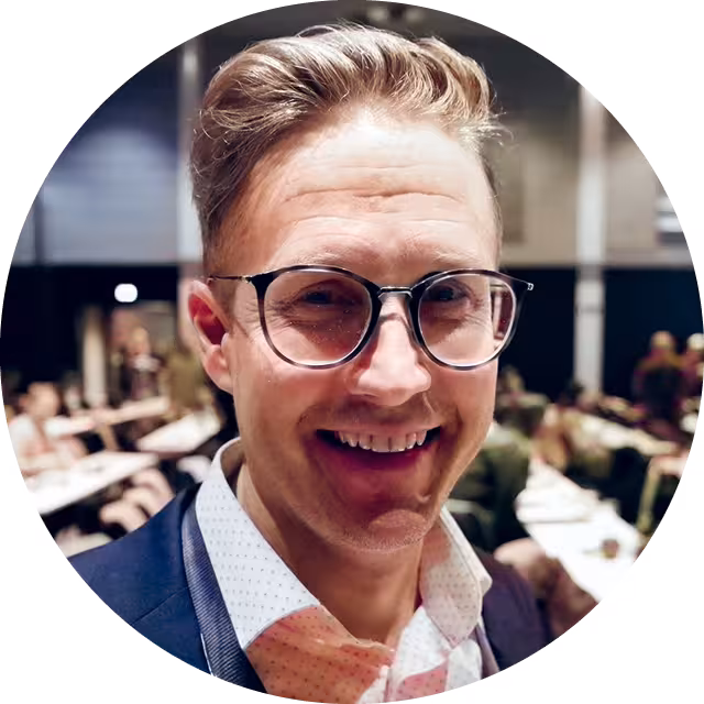 Portrettbilde av mann i skjorte og blazer, med briller, som står i konferansesal, smiler og ser inn i kameraet