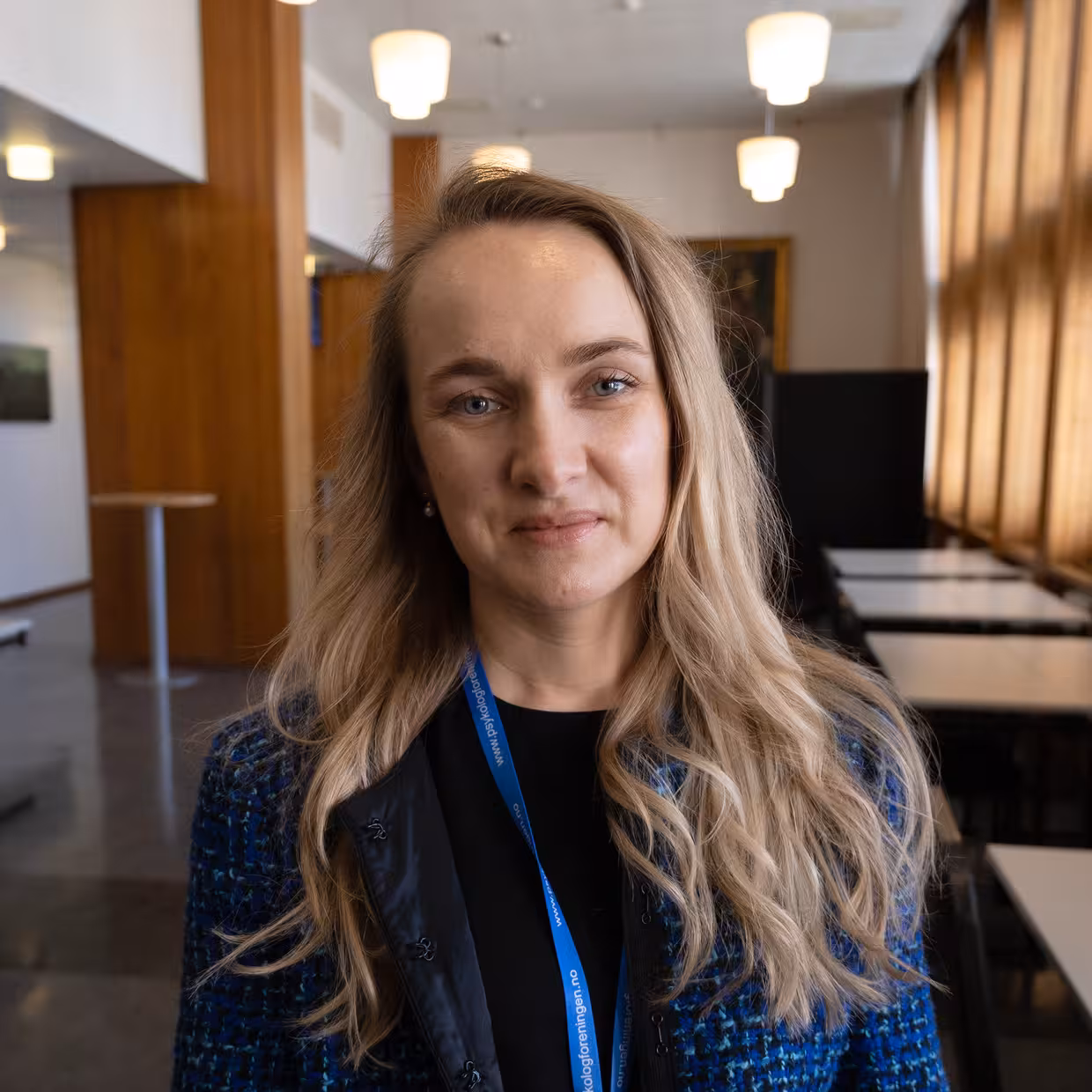 Nærportrett av en person med langt lyst hår som står i et rom med trepanel og taklamper; personen har på seg en mørk topp og en blå konferansesnor rundt halsen.