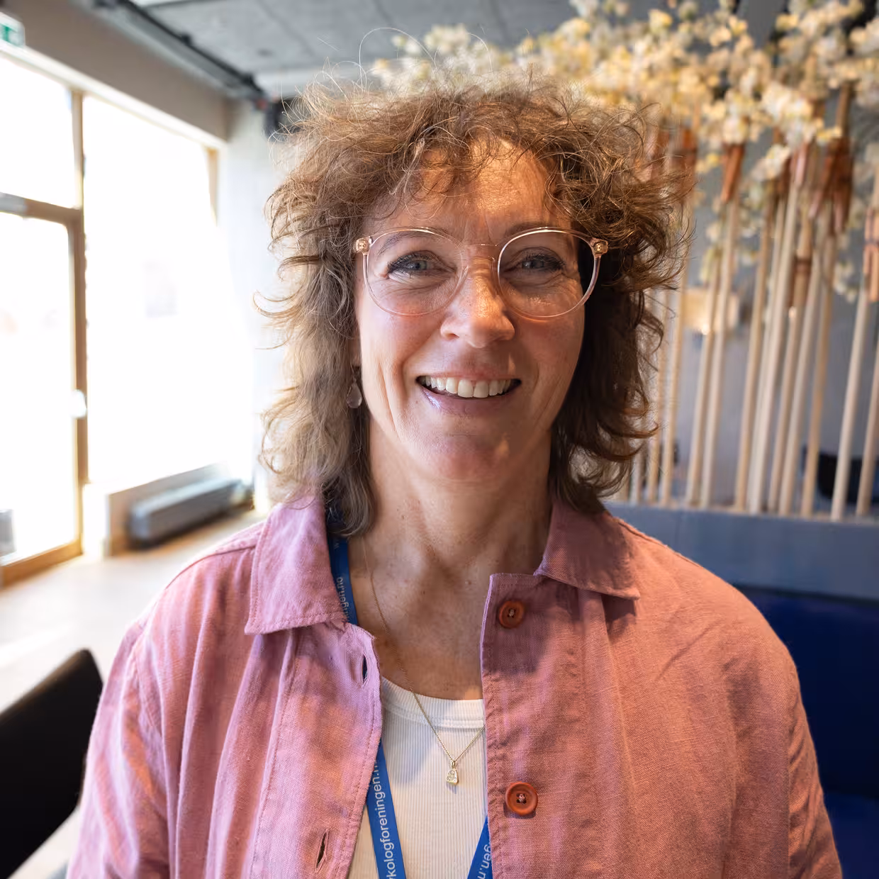 Nærportrett av en person med krøllete hår og runde briller, iført en rosa skjorte, som står foran en sofaseksjon og en stor blomsterdekorasjon; rommet er lyst med store vinduer og dekorative elementer.