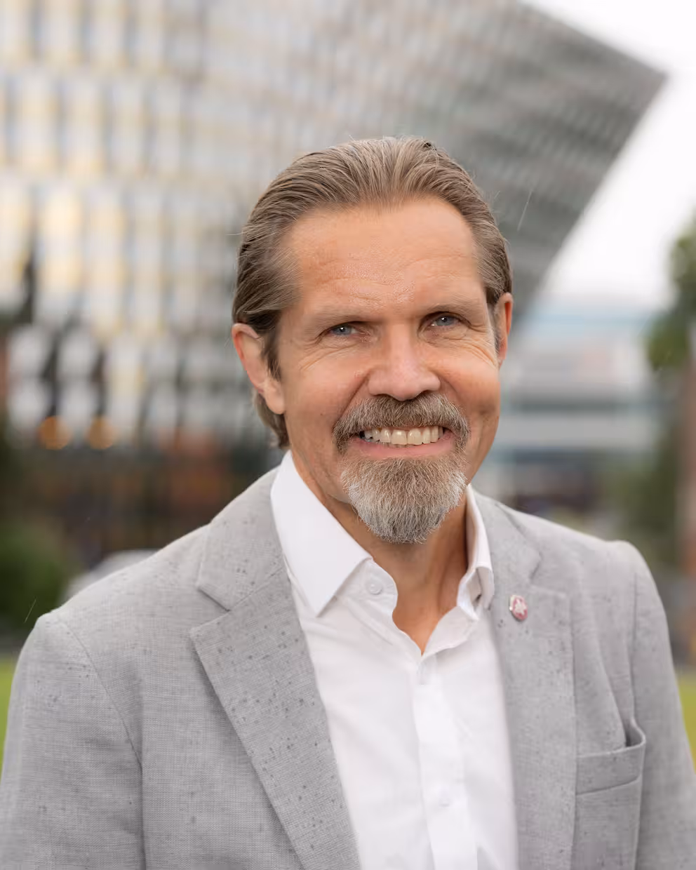 Portrett av en person i lys grå blazer og hvit skjorte, fotografert utendørs. I bakgrunnen sees en moderne bygning med glassfasade i uskarpt fokus, og det er synlige regndråper på jakken og i luften.