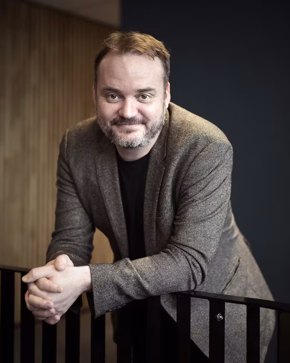 Portrett av en person i brun blazer og mørk genser, lent mot et svart rekkverk innendørs. Bakgrunnen består av en mørk vegg og vertikale trepaneler i mykt fokus.