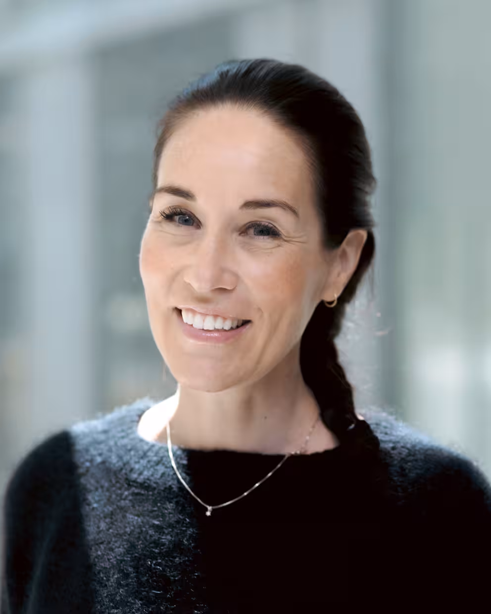 Portrett av en person med oppsatt hår og mørk skjorte, fotografert innendørs foran store vindusflater. Bakgrunnen er uskarp og består av lyse glass- og metallelementer.