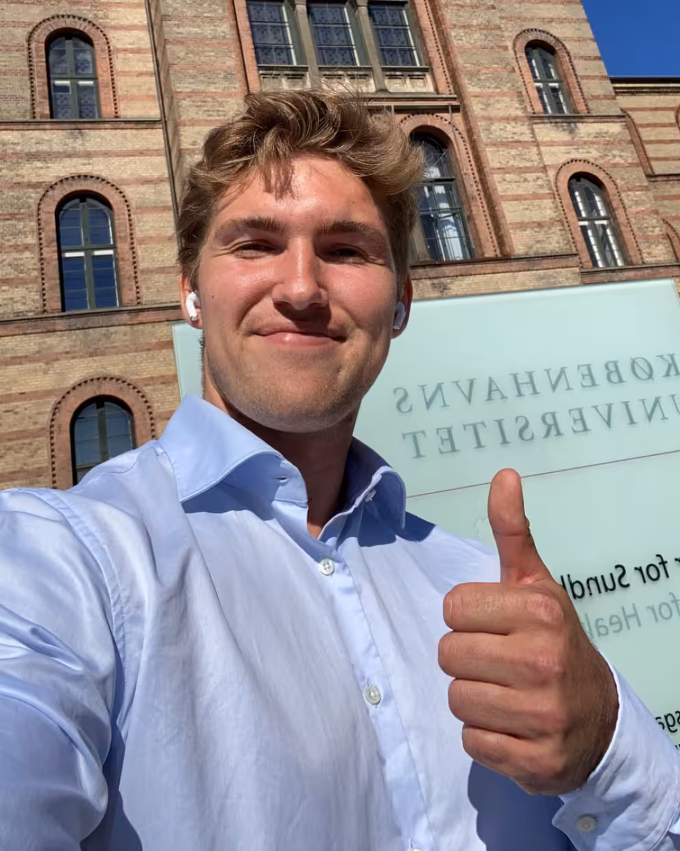 En person i lys skjorte tar en selfie foran en stor murbygning med kuppel og buede vinduer. Personen holder tommelen opp, og foran bygningen står et skilt med speilvendt tekst. På skiltet står det «Københavns universitet» i store bokstaver.