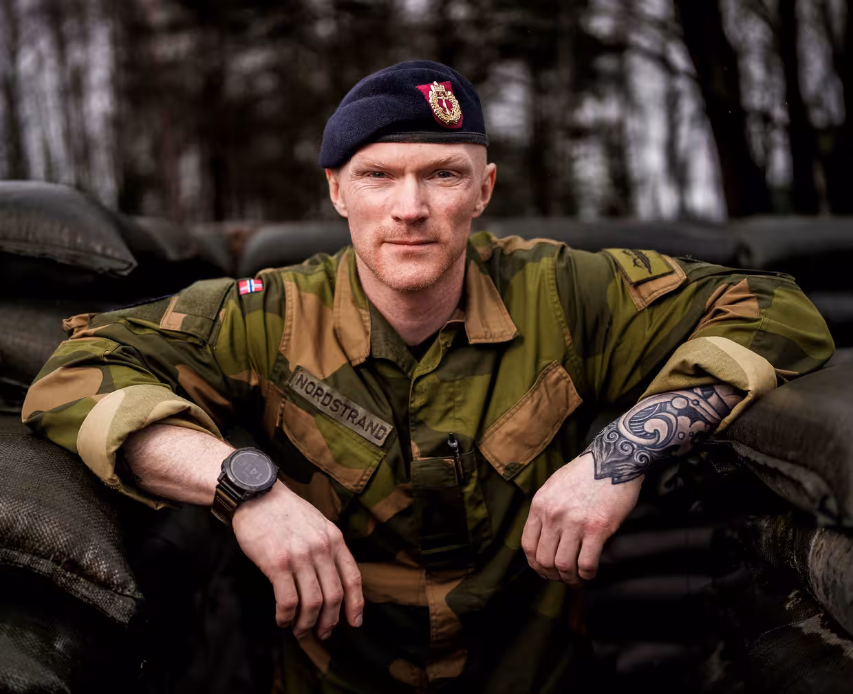 Portrett av en person i norsk militæruniform og mørk beret med emblem, lent med armene over mørke sandssekker utendørs. Personen har tatovering på underarmen og klokke på håndleddet, og bakgrunnen viser en skog med trær i dempet lys.