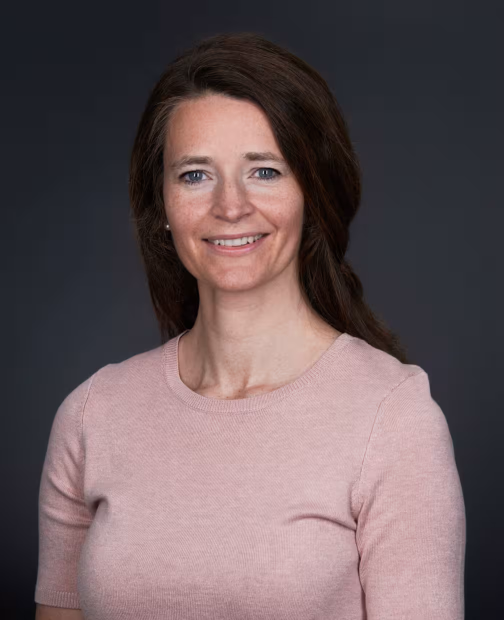 Studioportrett av en person med langt mørkt hår, iført en lys rosa kortermet genser, fotografert foran en mørk grå bakgrunn.