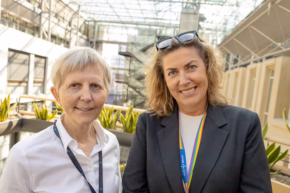 To personer står side om side i et lyst atrium med planter og glasskonstruksjoner i bakgrunnen. Begge har nøkkelbånd rundt halsen og er fotografert fra livet og opp.