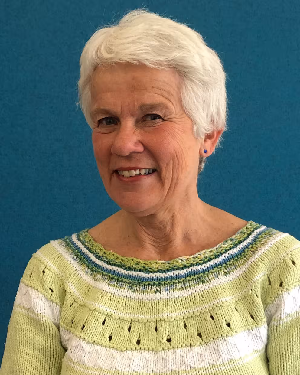 Portrett av en person med kort hvitt hår og lysegrønn strikkegenser, fotografert foran en ensfarget blå vegg. Personen er avbildet fra brystet og opp.