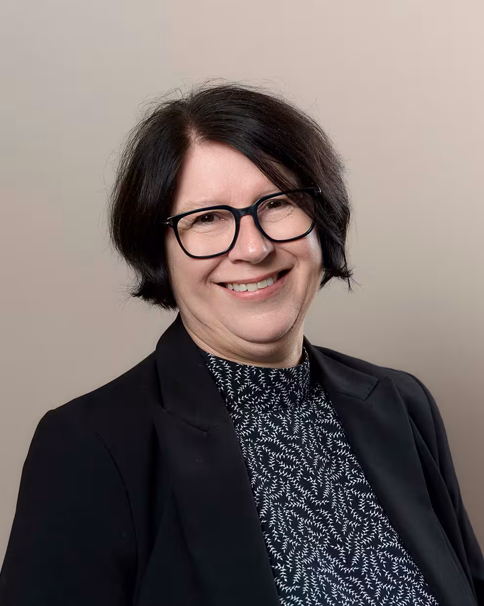 Portrett av en person med briller, mørk blazer og mønstret høyhalset overdel, fotografert mot en nøytral bakgrunn.